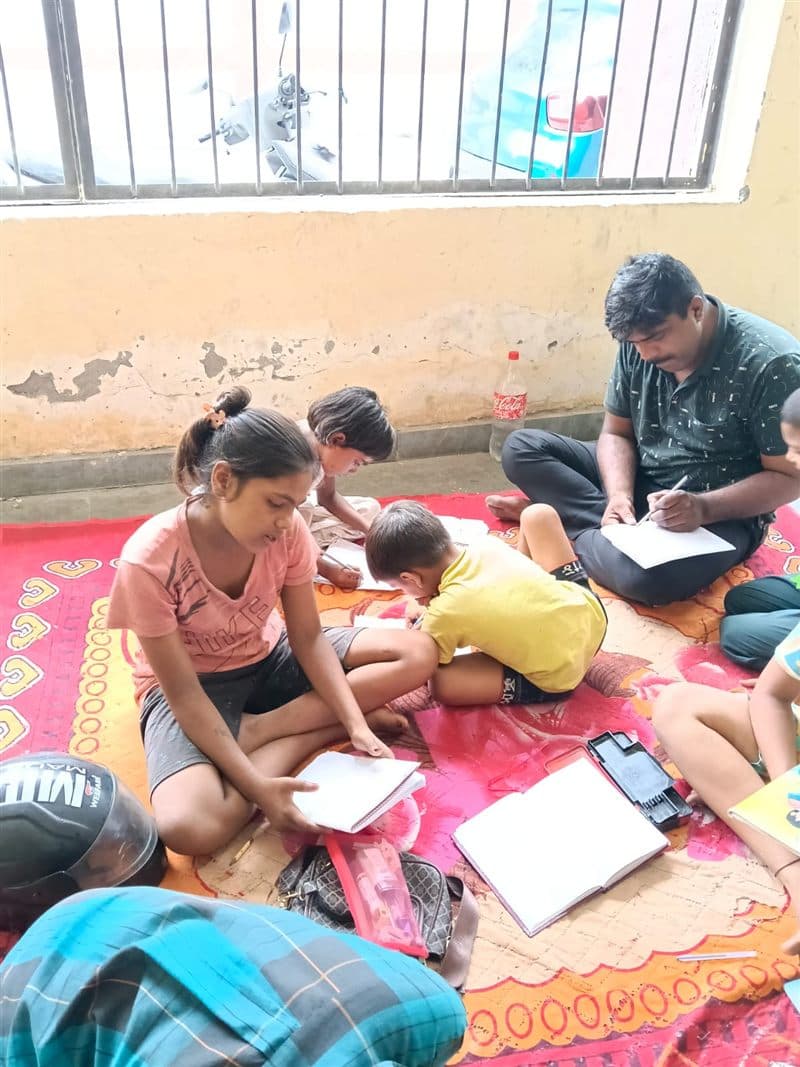 Children studying with notebooks on a mat while a tutor writes beside them