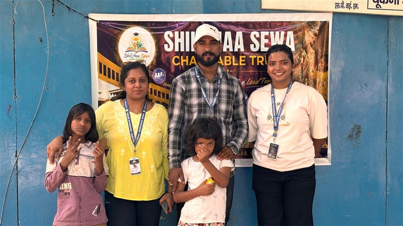 Three volunteers and two children standing in front of a Shikshaa Sewa Charitable Trust banner