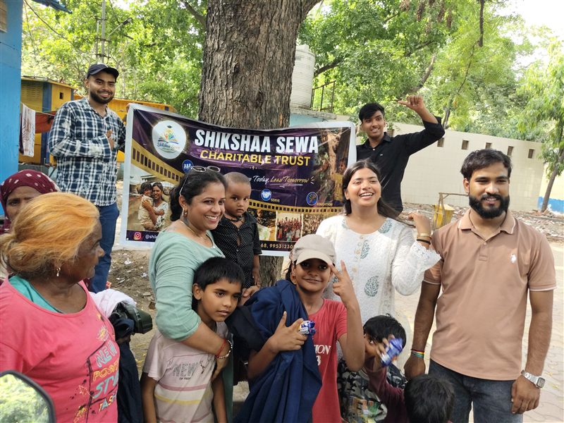 Volunteers and children gathered around a Shikshaa Sewa Charitable Trust banner under a tree